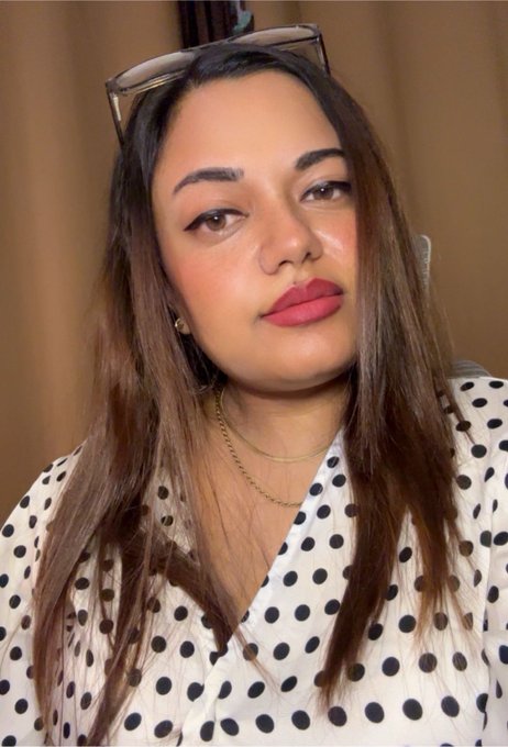 A woman with long brown hair, red lipstick, and sunglasses perched on her head wears a white blouse with black polka dots and layered gold chains.