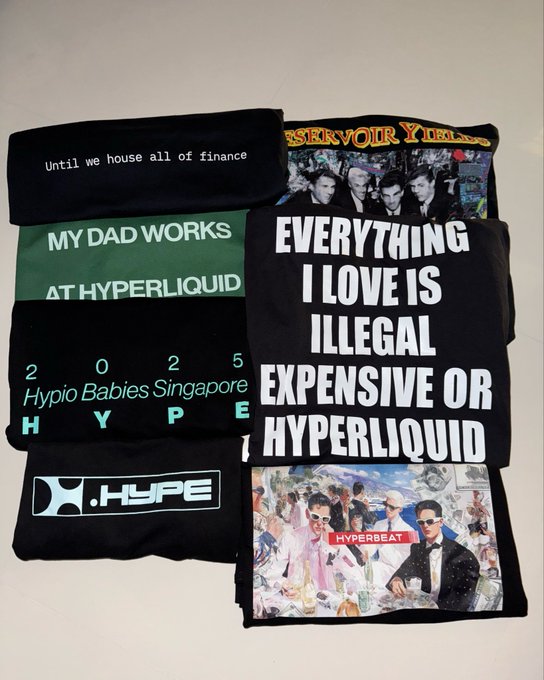A flat-lay collection of folded black t-shirts and apparel featuring Hyperliquid-related text and graphics, including phrases like 'MY DAD WORKS AT HYPERLIQUID', 'EVERYTHING I LOVE IS ILLEGAL EXPENSIVE OR HYPERLIQUID', and '2025 Hypio Babies Singapore HYPE'.
