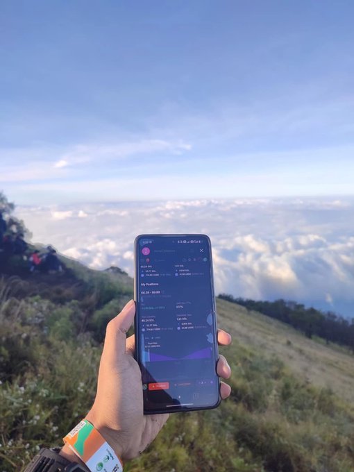 A hand holds a smartphone displaying a staking dashboard above a sea of clouds at a mountain summit, with "My Positions" visible on screen—perfectly matching the post's humorous checklist of "things you need to start LPing," where the only essentials are a mobile phone and internet connection, though a breathtaking view doesn't hurt.