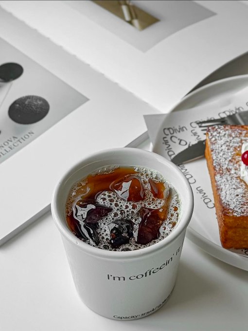 In the image, there are two cups of coffee placed next to each other on a table. One cup has a white lid that says "I'm Caffeine" written on it, while the other cup does not have any writing on its lid. The table also holds a book and a plate with a pastry on it.