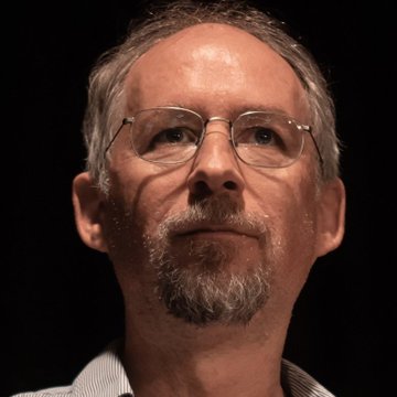 A close-up portrait photograph shows an older man with glasses, gray hair, and a beard against a dark background, resembling an academic or author portrait.