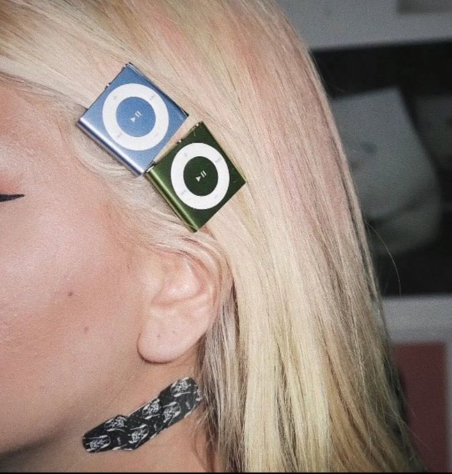 A close-up of a blonde woman wearing two iPod Nano hair clips (one blue, one green) in her hair, with a silver chain earring, against a dark background.