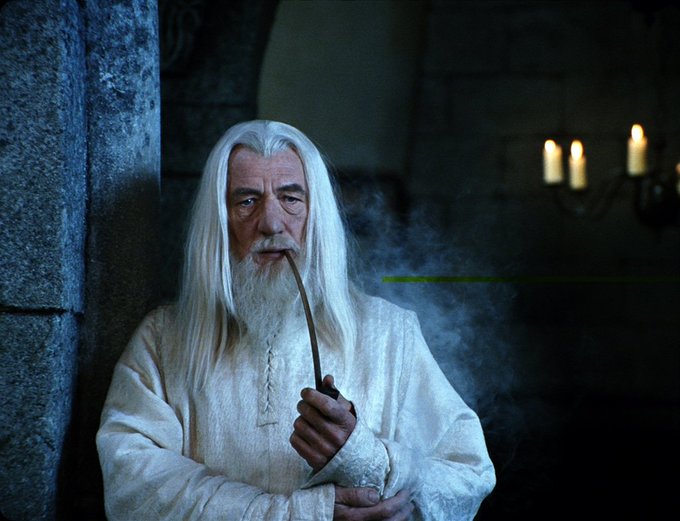 A man with long white hair and beard, resembling the character Gandalf, smokes a pipe in a dark stone setting with candles in the background.