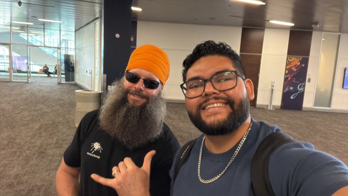Two men take a selfie in what appears to be an airport terminal; the man on the left wears an orange turban, sunglasses, and a beard, while the man on the right wears glasses and a blue shirt.
