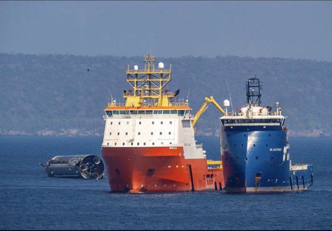 A photograph of two large offshore vessels—one red and white, the other blue—operating at sea near a forested coastline, with a submerged cylindrical object visible in the water.