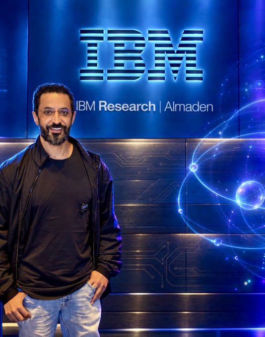 A man with a beard smiling in front of an illuminated 'IBM Research | Almaden' sign and a glowing blue digital globe display.