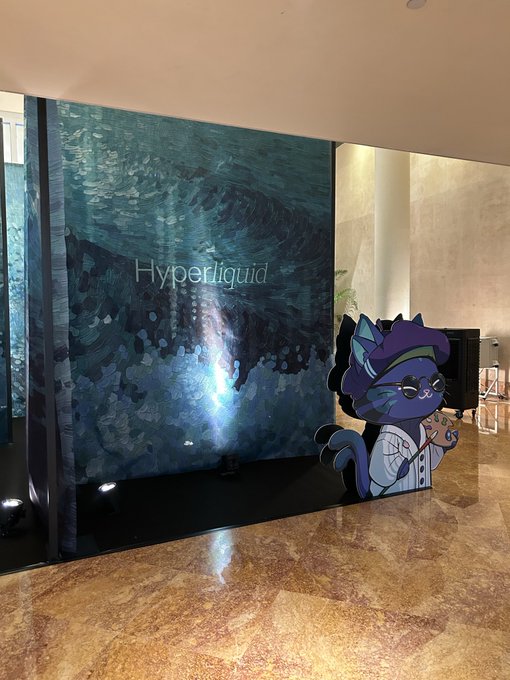 A plush cartoon cat cutout stands next to a large teal and blue textured display featuring the "Hyperliquid" logo in a sleek modern lobby, capturing the branded atmosphere of a Hyperliquid side event.