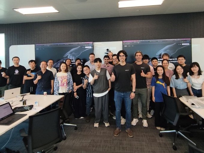 A group photo of around twenty people posing in a conference room with presentation screens in the background.