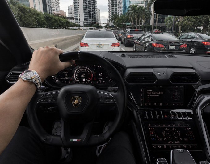 A first-person view from inside a Lamborghini, showing the driver's hand on the steering wheel (wearing a luxury watch) and the dashboard display, with traffic visible ahead on what appears to be a city highway.