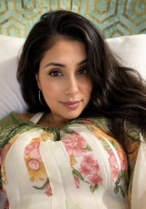 A selfie of a woman with long dark wavy hair wearing a floral-print dress with pink and orange flowers, posing against a geometric-patterned background.