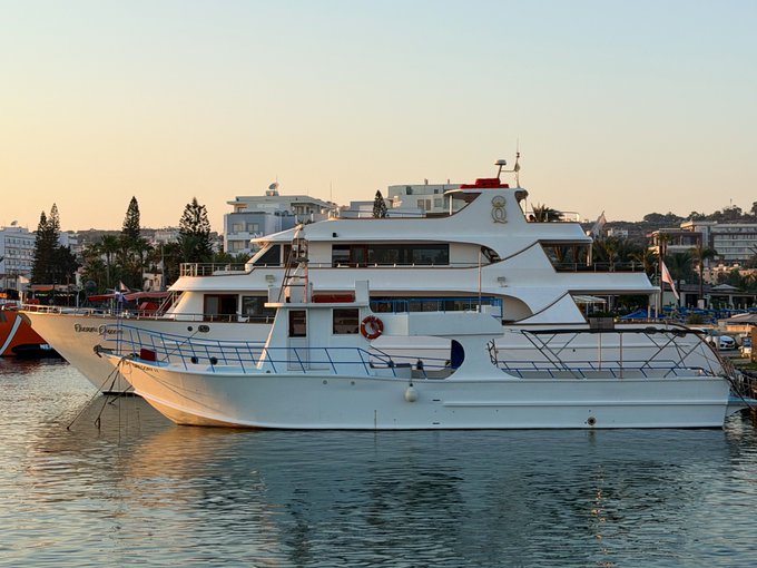 A white multi-deck yacht named "Dream Guardian" is docked in a harbor at dusk with a city skyline and palm trees in the background.