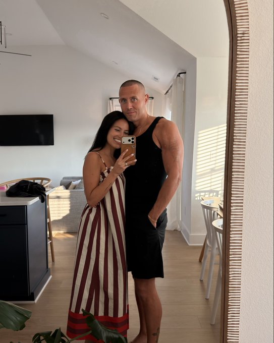 A couple takes a mirror selfie in their bright living room, smiling warmly as the woman in a striped sundress and the muscular man in a black tank top pose together with her phone. The casual, sunlit home setting complements the cheerful tone of celebrating "green candle day" together.