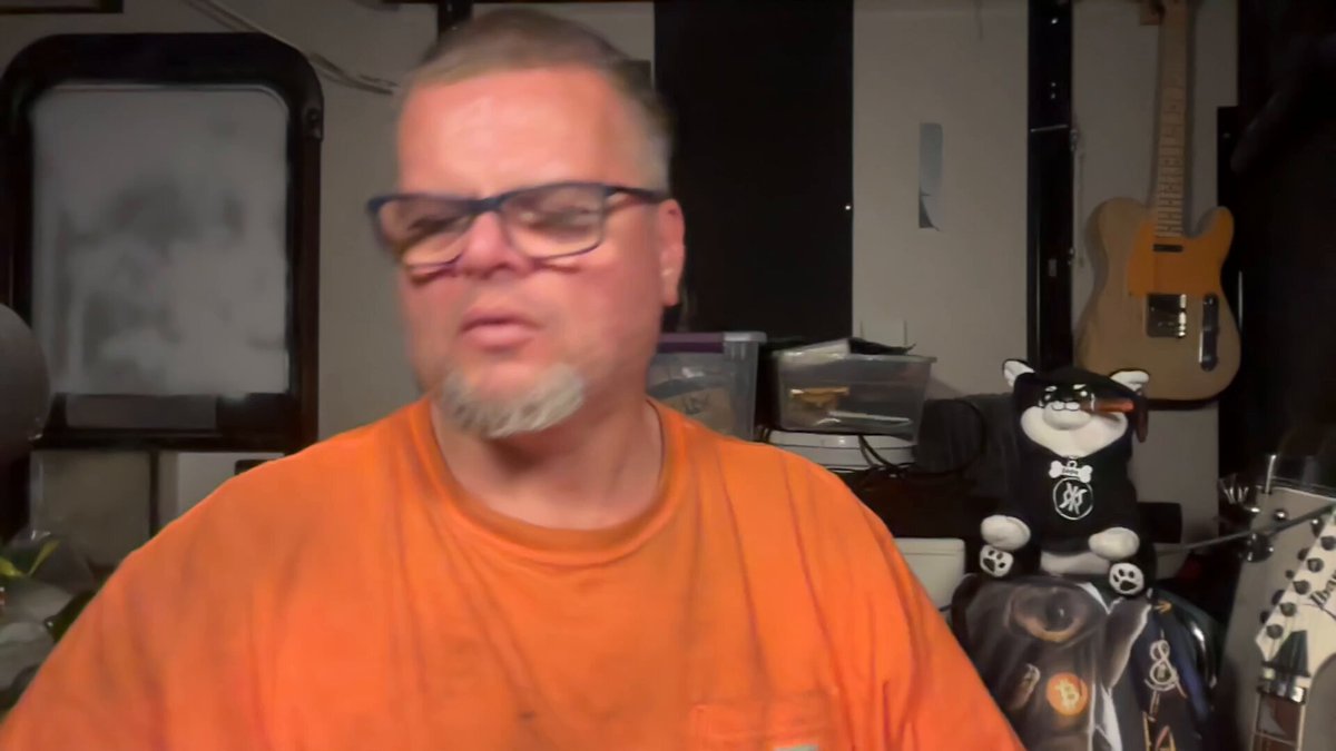 The image features a man wearing an orange shirt, sitting behind a desk with a guitar next to him. He has glasses on his face and appears to be talking or posing for a photo. The background includes a wall clock and a poster of a cat. There is also a text overlay in the image that reads "Buy the bitcoin blockchain #1 $DOG".
