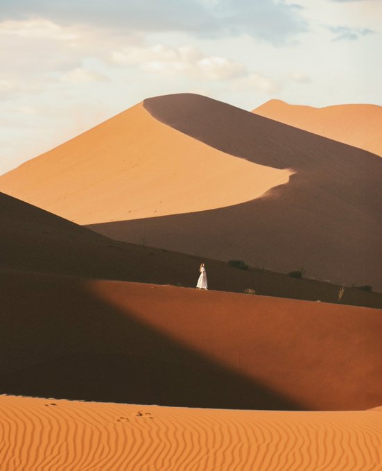 A person in a white robe stands on a vast desert dune, dwarfed by towering sand dunes in warm orange tones under a cloudy sky.