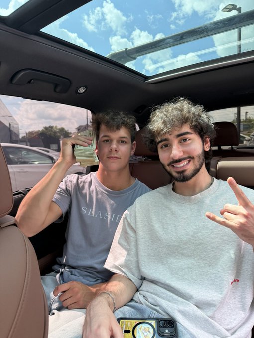 Two young men sit inside a car with a sunroof, smiling at the camera while one holds up a stack of cash and the other makes a hand gesture, with visible text "SEASIDE" on one shirt. The image relates to the post by visually suggesting the promise of real-world "airdrops" of money, as $ANSEM is known for a meme culture of giving cash to fans in person.
