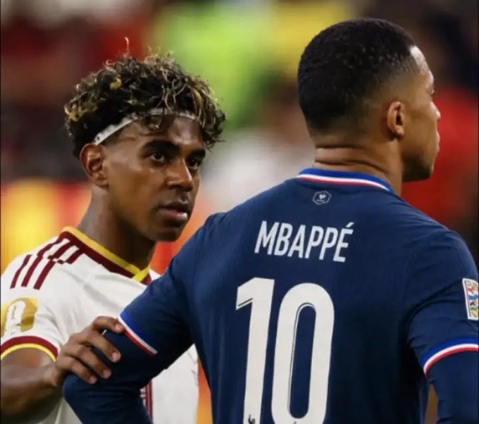 Two soccer players; one in a white jersey looking at the back of another player wearing a dark blue France national team jersey with 'MBAPPÉ' and the number '10'.