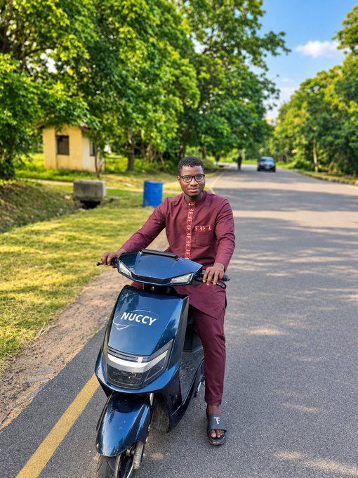 A man in a maroon traditional outfit stands next to a dark blue scooter labeled 'NUCCY' on a tree-lined street.