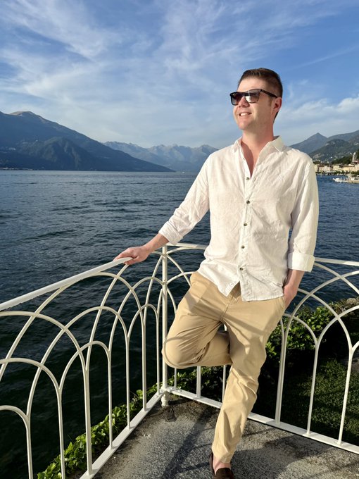 A photograph of a smiling man wearing sunglasses, a white button-down shirt, and khaki pants, leaning against a white metal railing by a body of water with mountains in the background.