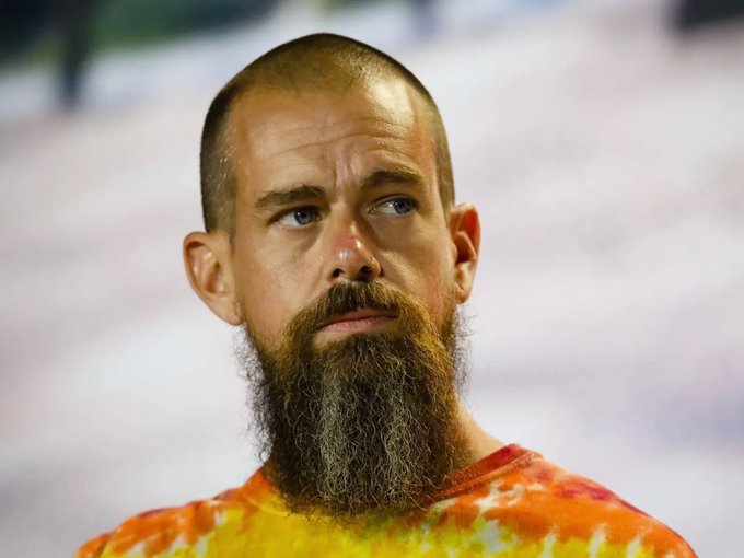 A close-up portrait of a bald man with a long, full beard wearing an orange and yellow tie-dye shirt, with what appears to be a small amount of blood on his nose, looking off to the side.