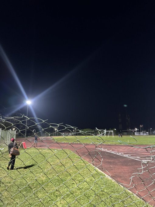 A nighttime view through a torn chain-link fence at a stadium track, illuminated by bright stadium lights, with the "WAUG." post likely referring to the West Africa University Games or a similar event held at the athletic facility.