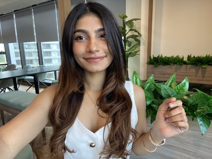A young woman with long brown hair wearing a white top and delicate necklace smiles at the camera in what appears to be a modern office or apartment setting with plants and furniture in the background.