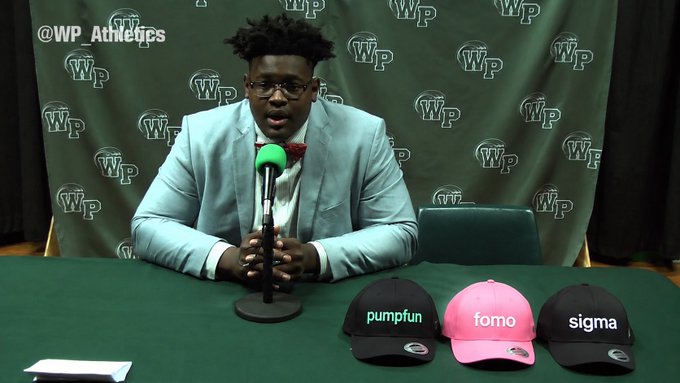 A man in a light blue blazer and glasses sits at a table with three baseball caps labeled "pumpfun," "fomo," and "sigma," preparing to reveal his choice in front of a William Paterson (WP) Athletics backdrop. The setup mirrors the "tonight I reveal" theme, teasing an announcement or selection among the three caps.