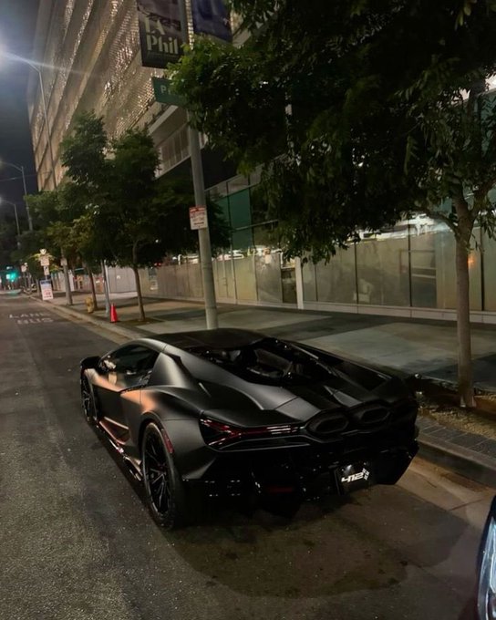A matte black sports car is parked on a city street at night next to a sidewalk with trees and a building with a 'Philz' sign.