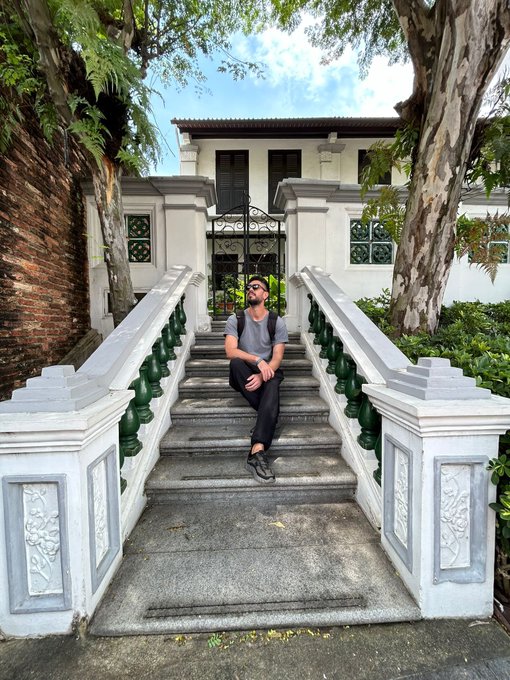 A man wearing sunglasses sits on stone stairs leading to a white building with an ornate gate, surrounded by greenery.