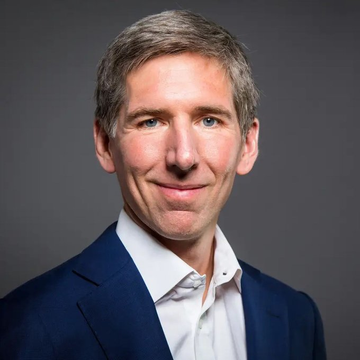 A professional headshot of a middle-aged man with short gray hair, wearing a dark blue suit jacket over a white shirt, smiling slightly against a gray background.