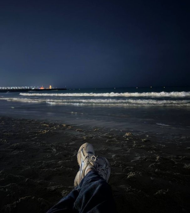 A first-person view of feet resting on a sandy beach at night, looking out at gentle waves and a distant, illuminated city skyline.