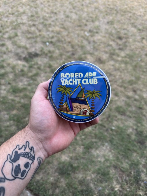 A hand with a cartoon cat tattoo holds a circular sticker or coaster labeled "BORED APE YACHT CLUB" featuring palm trees and a sailboat design, set against a grassy outdoor background.