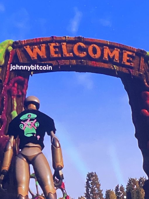 A robot wearing a black t-shirt featuring a green Bored Ape logo stands beneath a "WELCOME" archway, with the username "johnnybitcoin" visible.