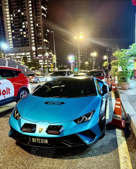 A blue Lamborghini is parked on a city street at night with a license plate reading 'BITCOIN'.