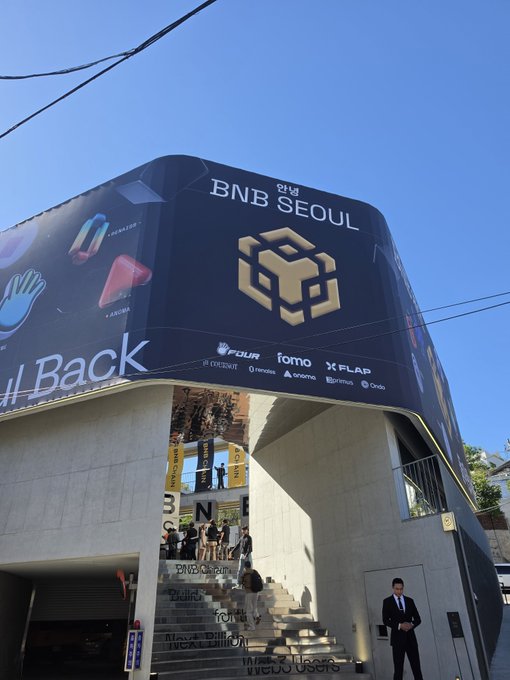An exterior shot of a building with a large black billboard for 'BNB SEOUL' featuring the BNB chain logo, alongside other Web3 sponsors like Four, Fomo, and X FLAP.