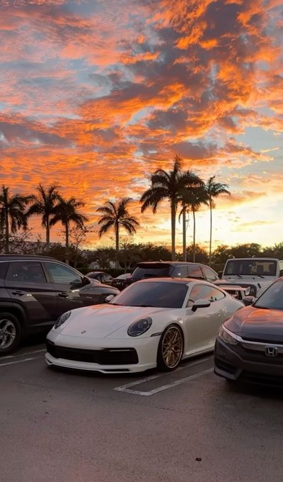 A scenic photograph of a parking lot at sunset featuring a white Porsche 911 in the center, flanked by a black SUV and a dark Honda, set against a dramatic orange and pink cloudy sky with silhouetted palm trees.