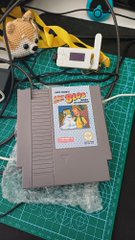 A Nintendo Entertainment System (NES) game cartridge with a colorful sticker on it sits in bubble wrap on a green cutting mat, with a hot glue gun and other crafting tools visible in the background.