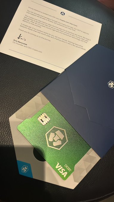The image shows a green Visa card placed inside an envelope, which is sitting on top of a table. There are also two pieces of paper present in the scene - one is a letter addressed to someone named "Kris" written on a piece of white paper and the other is a note with black text that reads: "It has arrived!!!! Thank you @cryptocom". The envelope appears to be opened, revealing the contents inside.