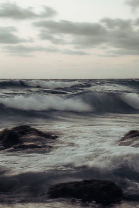 A photograph captures dark, turbulent ocean waves rolling under an overcast, grey sky, with dark rocks visible in the foreground.