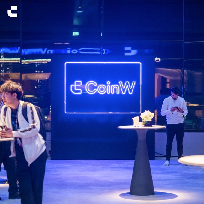Two men stand near a tall cocktail table with a floral centerpiece, while a large illuminated blue sign displaying the "CoinW" logo glows prominently in the background, suggesting a promotional or networking event for the cryptocurrency exchange platform.
