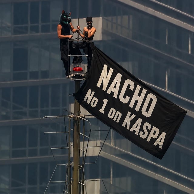 Two people wearing masks stand on a tall pole structure, with one holding a black flag that reads 'NACHO No 1 on KASPA' in white letters.