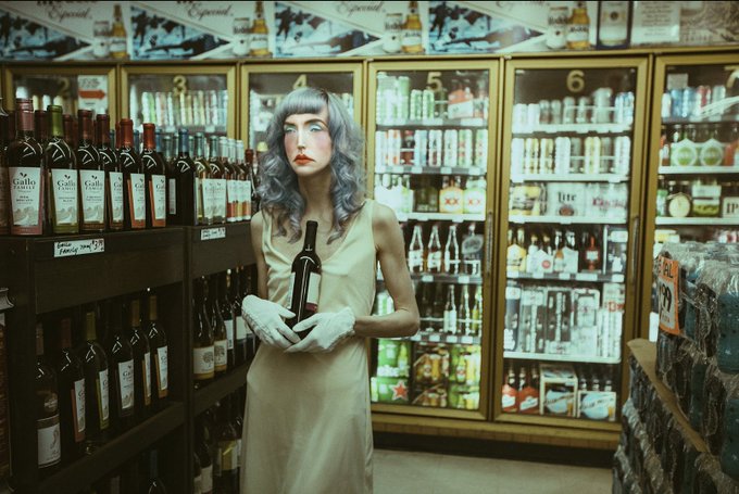 A striking portrait of a pale, silver-haired woman in a cream-colored dress and white gloves holding a wine bottle inside a liquor store aisle, with numbered refrigerated sections (1-7) and shelves stocked with various wines and spirits visible in the background. The opensea.io reference suggests this is an NFT artwork titled "Los Angeles Portrait" created in 2017, capturing the uncanny, stylized aesthetic the platform is known for showcasing.