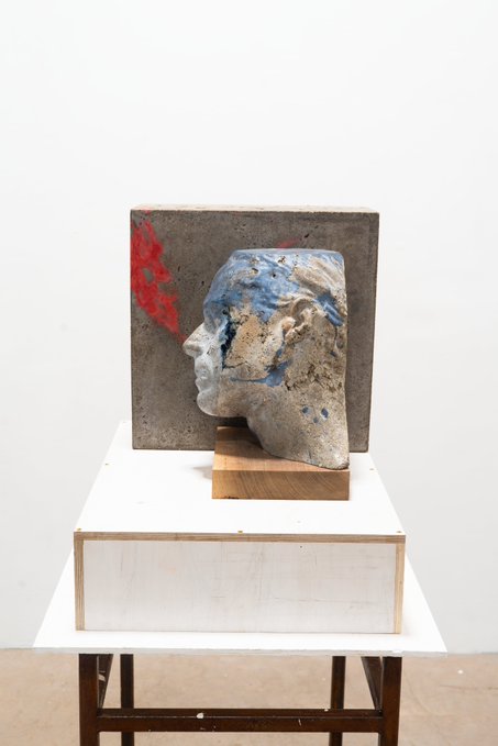 A mixed-media sculpture depicting a fragmented gray head in profile mounted on a concrete block, placed on a white plinth in a gallery setting.