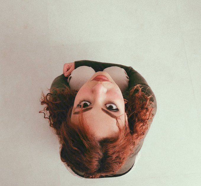 An upside-down artistic photo of a woman with curly red hair and red lipstick wearing a white top, set against a plain light background.