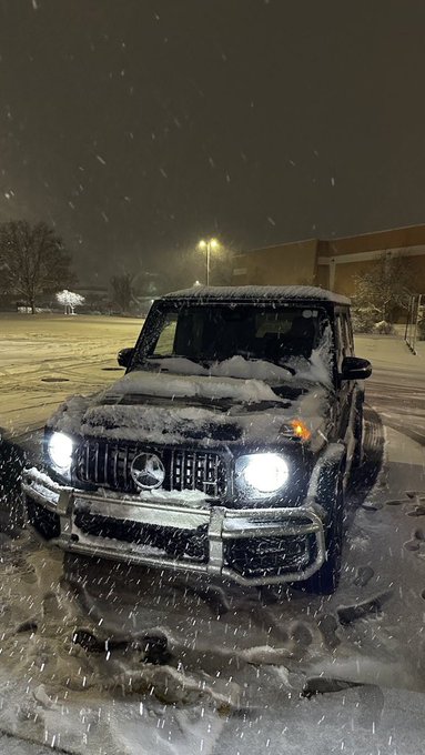 A Mercedes-Benz SUV is parked in a snowy parking lot, with snow falling around it. The car has its headlights on, illuminating the scene. In the background, there are buildings visible through the falling snowflakes.