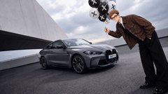 A man in a brown jacket holds up what appears to be a camera rig, posing next to a dark-colored Lexus sedan parked against a modern building.