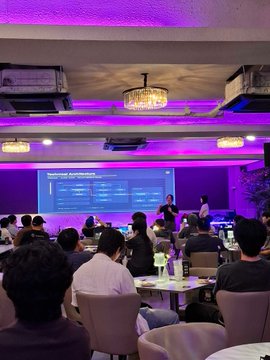 An indoor event or conference room with purple and pink lighting, featuring a chandelier, air conditioning units, and attendees seated at tables facing a presentation screen displaying technical information.
