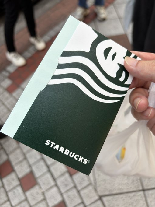 A hand holding a Starbucks paper bag with the iconic green siren logo while standing on a brick-tiled street.