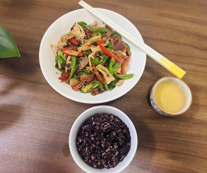 A wooden table set with a white bowl of stir-fried chicken and green peppers with chopsticks, a smaller bowl of black rice, and a small cup of yellow tea.