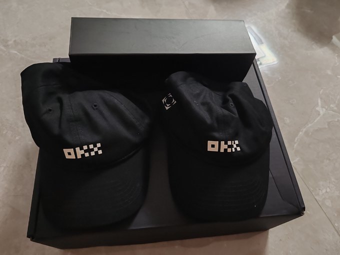 Two black baseball caps with the 'OKX' logo in white, displayed on a black box on a marble surface.