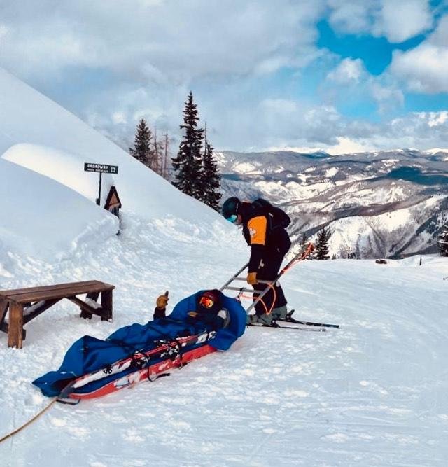 A person in ski gear and a helmet pulls a rescue sled carrying a patient in a blue bag across a snowy mountain landscape. A 'DEER CREEK' trail sign, picnic table, and pine trees are visible nearby.
